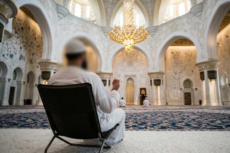 A man sitting on a chair inside a modern mosque, illustrating the context of the Fiqh ruling on praying while seated due to medical necessity.
